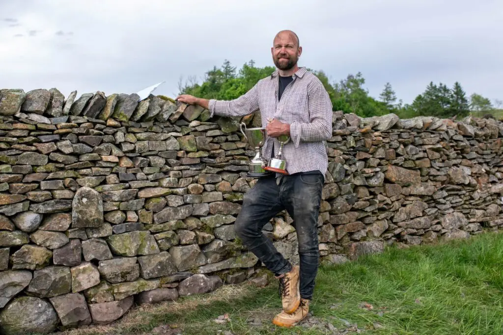 A man holding two trophies near a dry stone wall