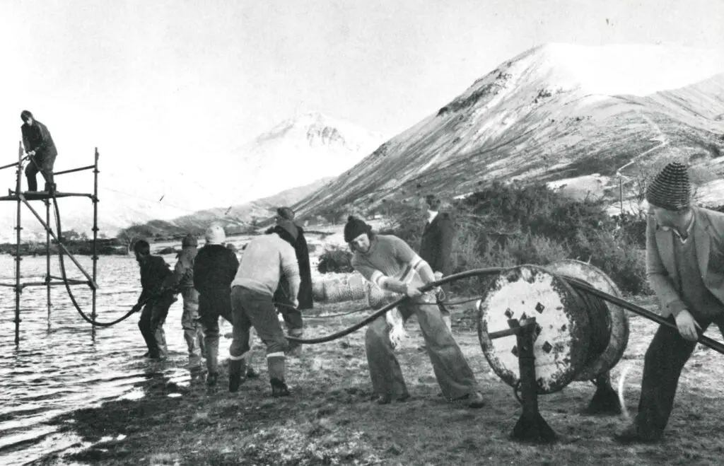 An old photo of people puling a cable and laying it under a lake