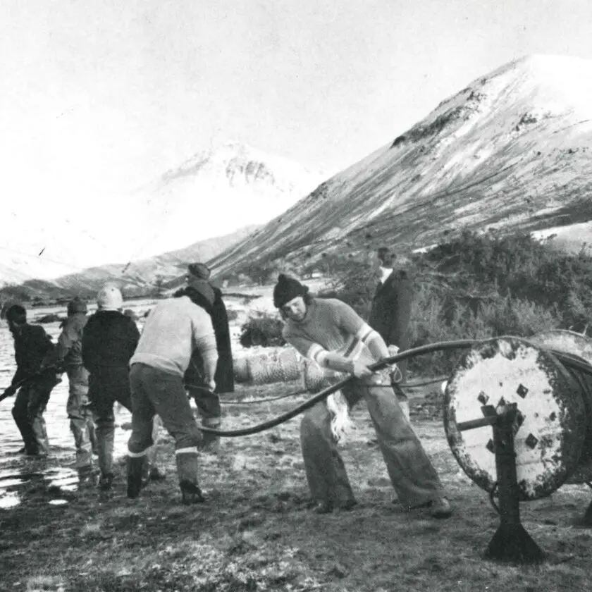 An old photo of people puling a cable and laying it under a lake