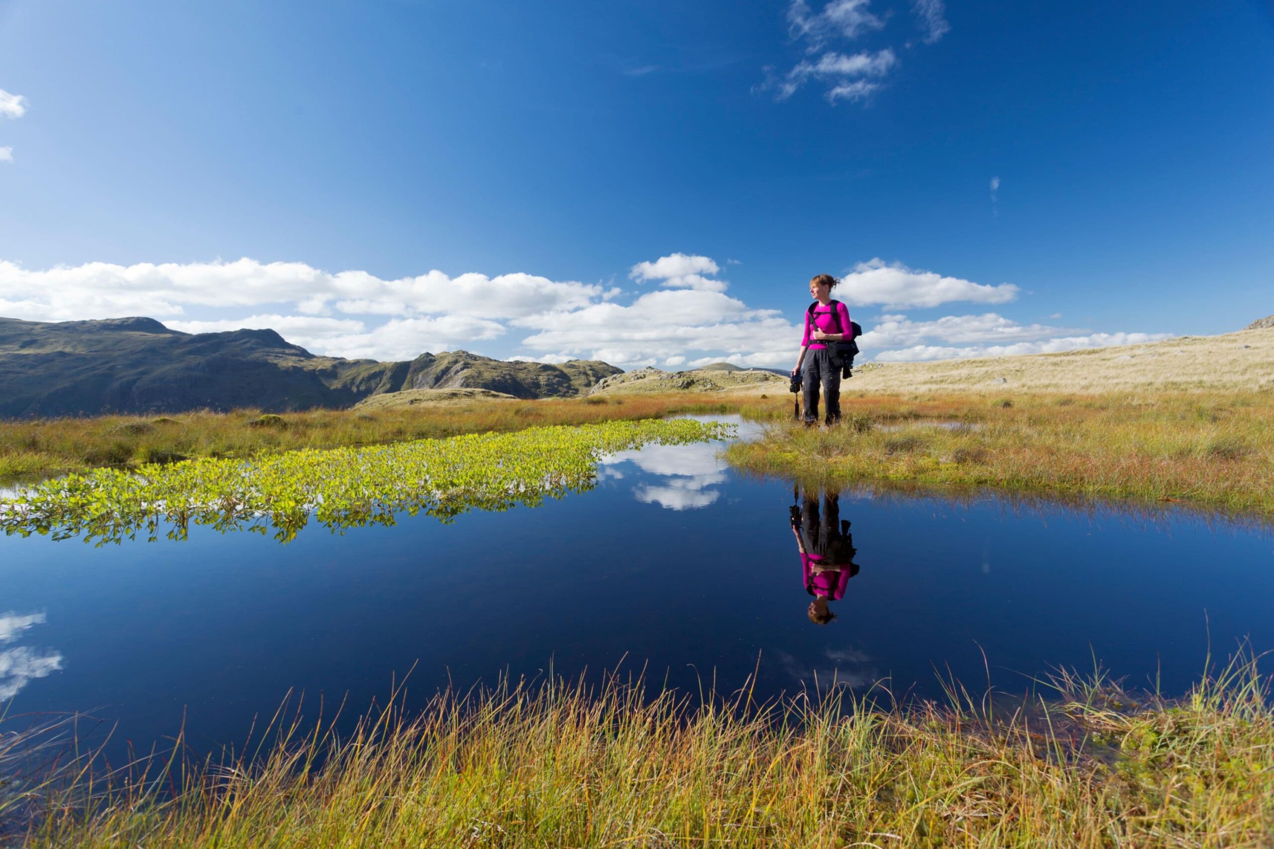 A woman stands at the edge of a tarn on a sunny day